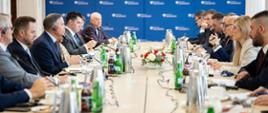 Wiceminister Robert Kropiwnicki wraz z przedstawicielami MAP, siedzą przy stole podczas spotkania z przedstawicielami Amerykańskiego przemysłu obronnego. Na stole dzbanki, butelki z wodą, słuchawki, mikrofony oraz kwiaty. W tle ścianka MAP