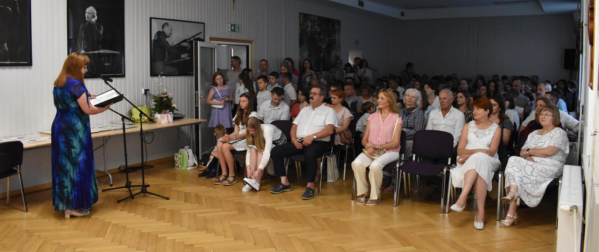 W prawej stronie zdjęcia Pani Dyrektor przemawia do publiczności w auli Państwowej Szkoły Muzycznej. Dół zdjęcia w brązowym kolorze, a górna część zdjęcia w odcieniach koloru białego. Większa część fotografii przedstawia osoby uczestniczące w wydarzeniu.