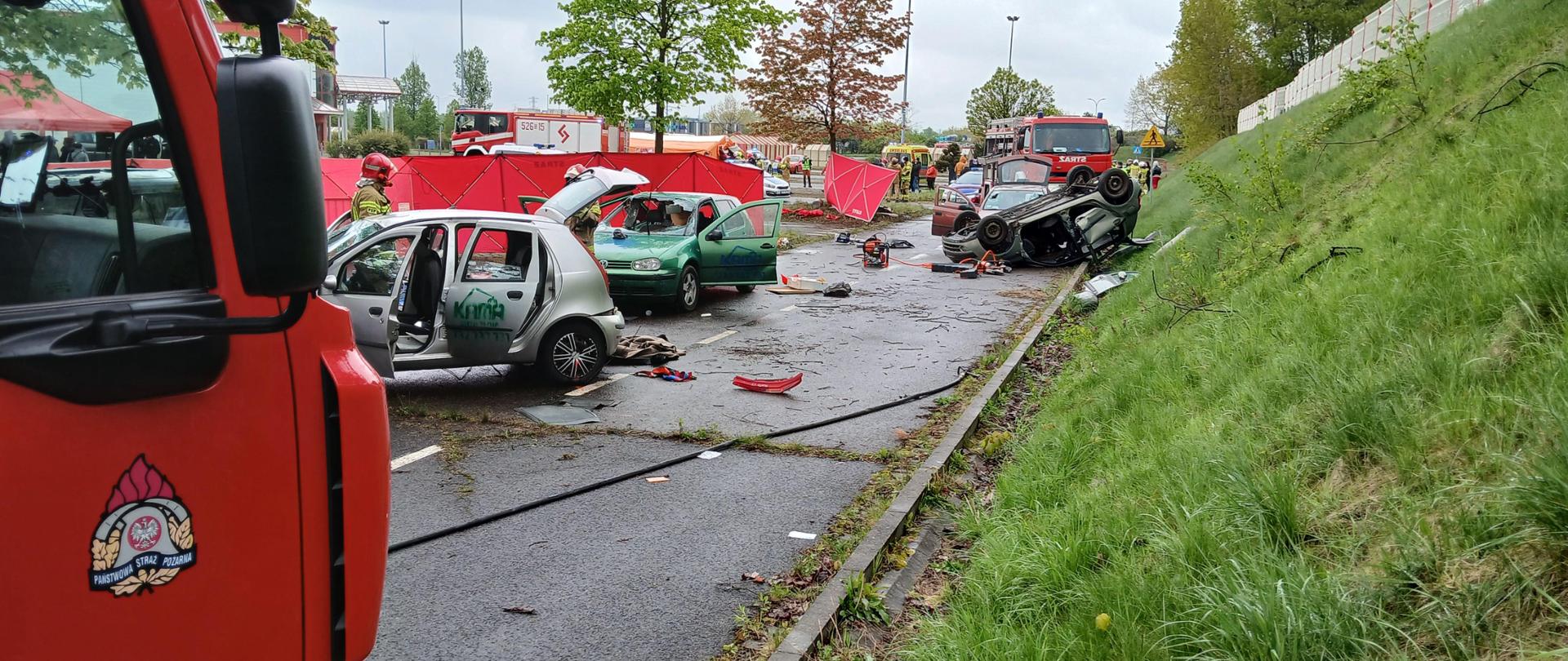 na drodze rozbite samochody osobowe z otwartymi drzwiami w tle strażacy i samochody strażackie podczas udzielania pomocy poszkodowanym w wypadku masowym. 