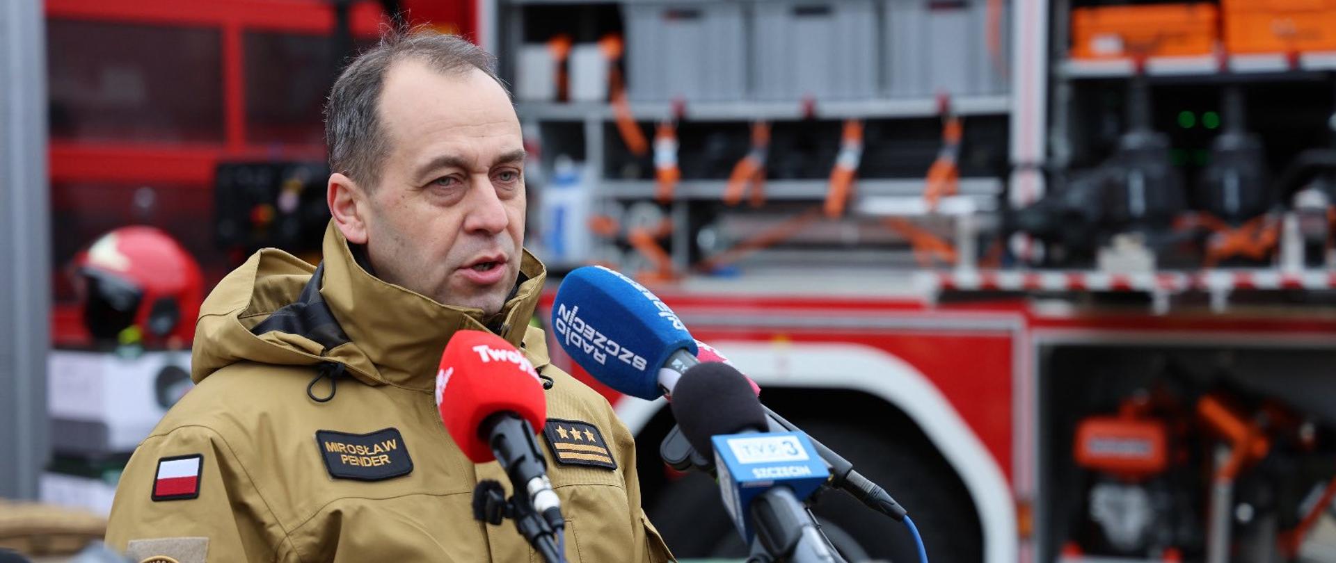 Strażak w mundurze przemawia do mikrofonów. W tle widoczny jest czerwony wóz strażacki z otwartymi schowkami na sprzęt.