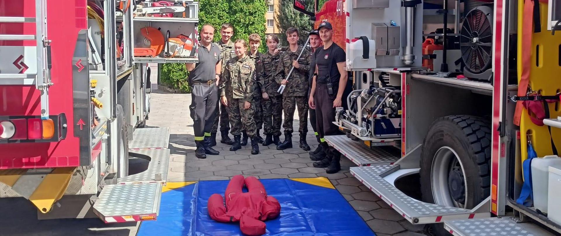 Na zdjęciu widać strażaków oraz grupę dzieci w mundurach moro, którzy między czerwonymi strażackimi samochodami pozują do zdjęcia. Samochody maja otwarte skrytki ze sprzętem. Przed nimi na betonie leży niebeska plandeka z żółtymi rogami, na której leży czerwony manekin. W tle widać bloki oraz krzewy. Jest widno.