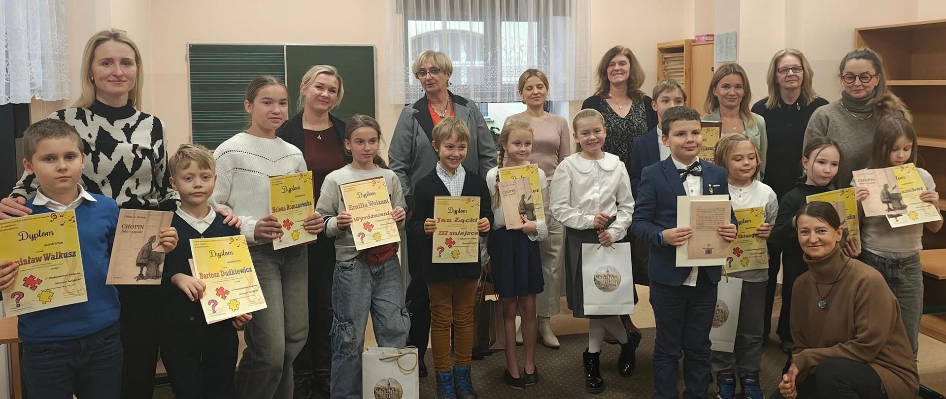 Do zdjęcia pozują uczestnicy konkursu trzymając dyplomy. Za nimi stoją nauczyciele.