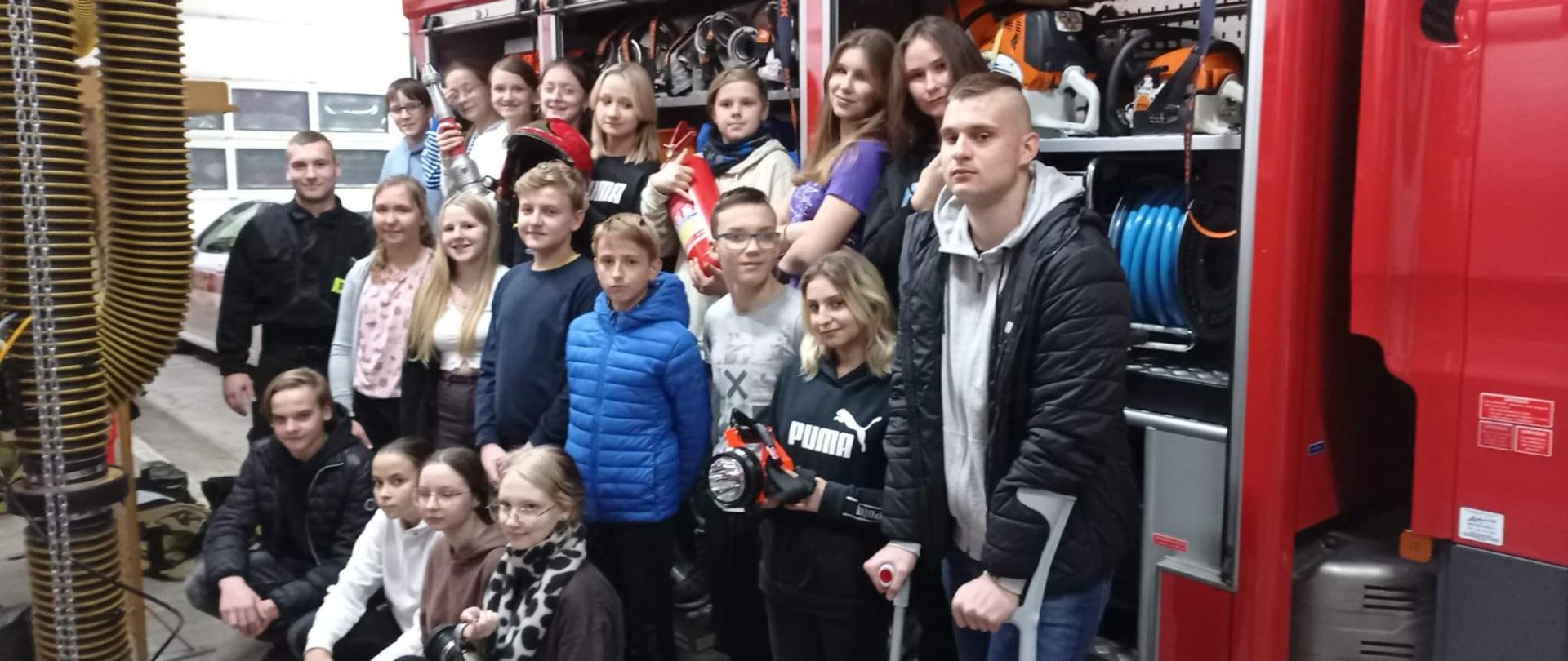Przed strażackim wozem bojowym w trzech rzędach stoi kilkanaście nastolatków.