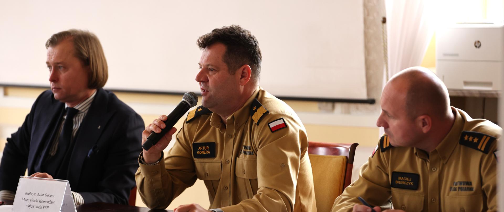 Wojewoda Mazowiecki w garniturze, Mazowiecki Komendant Wojewódzki PSP oraz Zastępca MKW PSP w piaskowych mundurach służbowych siedzą za stołem prezydialnym na beżowych krzesłach. Przed komendantami stoją tabliczki z nazwiskiem i funkcją. 