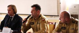 Wojewoda Mazowiecki w garniturze, Mazowiecki Komendant Wojewódzki PSP oraz Zastępca MKW PSP w piaskowych mundurach służbowych siedzą za stołem prezydialnym na beżowych krzesłach. Przed komendantami stoją tabliczki z nazwiskiem i funkcją. 