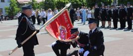 Poczet sztandarowy OSP w Lipnie przejmuje sztandar z rąk dh Prezesa. panowie klęczą na dywanie, sztandarowy całuje płat sztandaru. W tle zaloga w szeregu, za nimi widać orkiestrę. Plac apelowy w Lipnie.