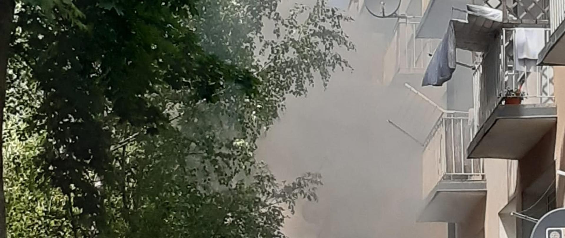 Zdjęcie przedstawia budynek mieszkalny wielorodzinny. Przed budynkiem widać dym. 