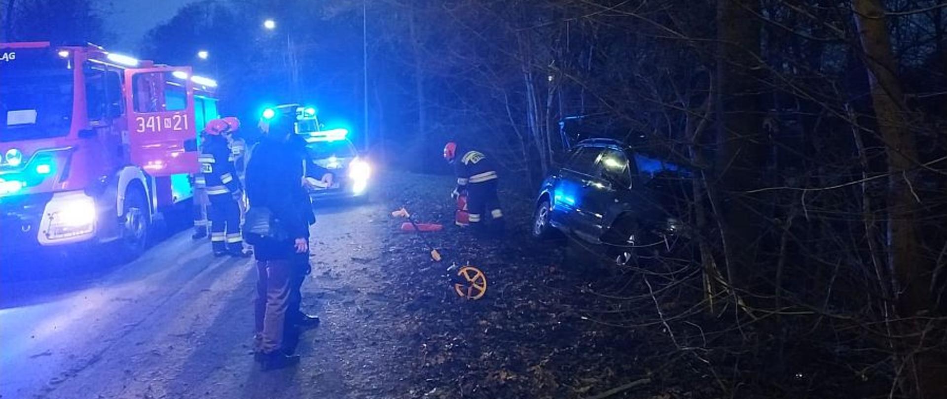 Po lewej stronie widoczny pojazd straży pożarnej stojący na skraju drogi. Po prawej stronie na poboczu pojazdy który uderzył w przydrożne drzewo, w środku stoją strażacy i policjant.