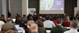 Sala konferencyjna. Uczestnicy siedzą przy stołach i słuchają kobiety, która przemawia stojąc za mównicą. Na ekranie widoczna jest prezentacja dotycząca ochrony torfowisk. W tle widoczne są banery Konwencji Ramsarskiej.