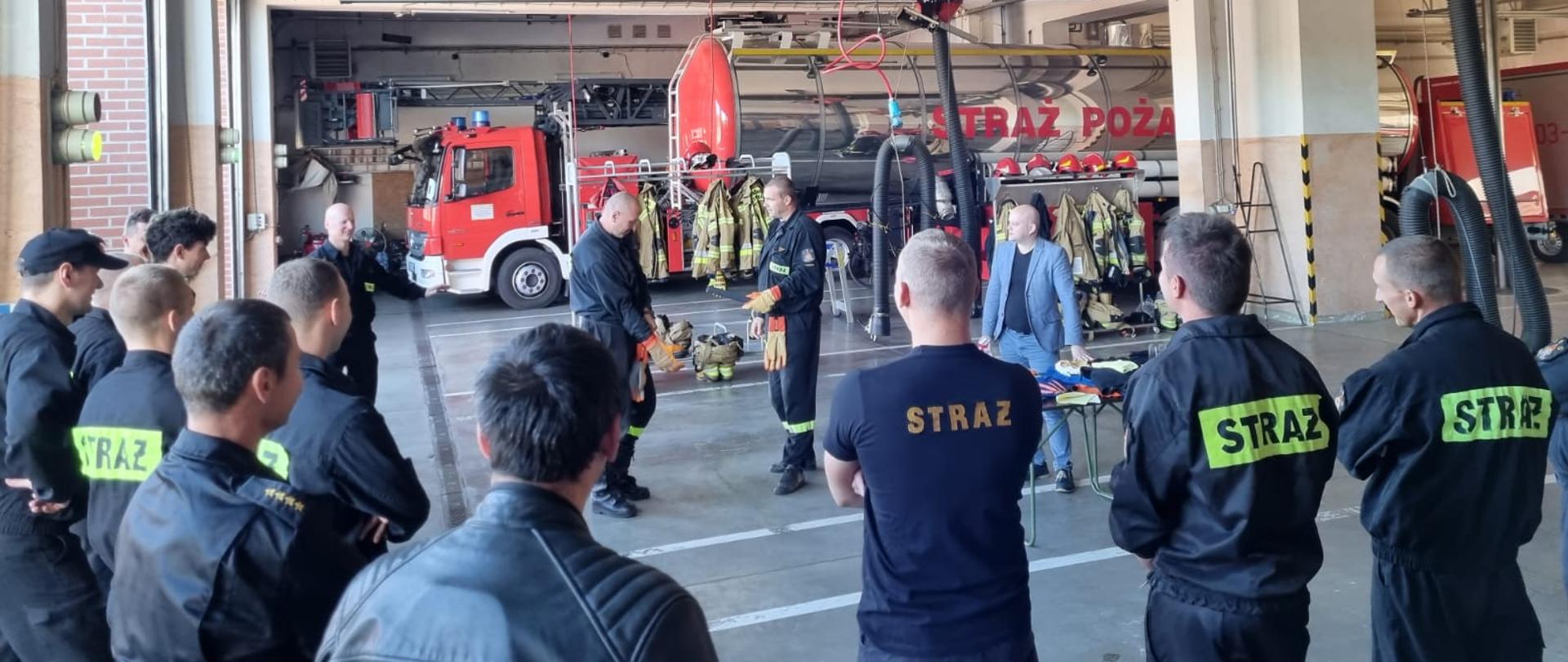 Uczestnicy pokazów stoją w garażu JRG3, w centralnej części zdjęcia przedstawiciel firmy i dwaj strażacy przymierzający rękawice.