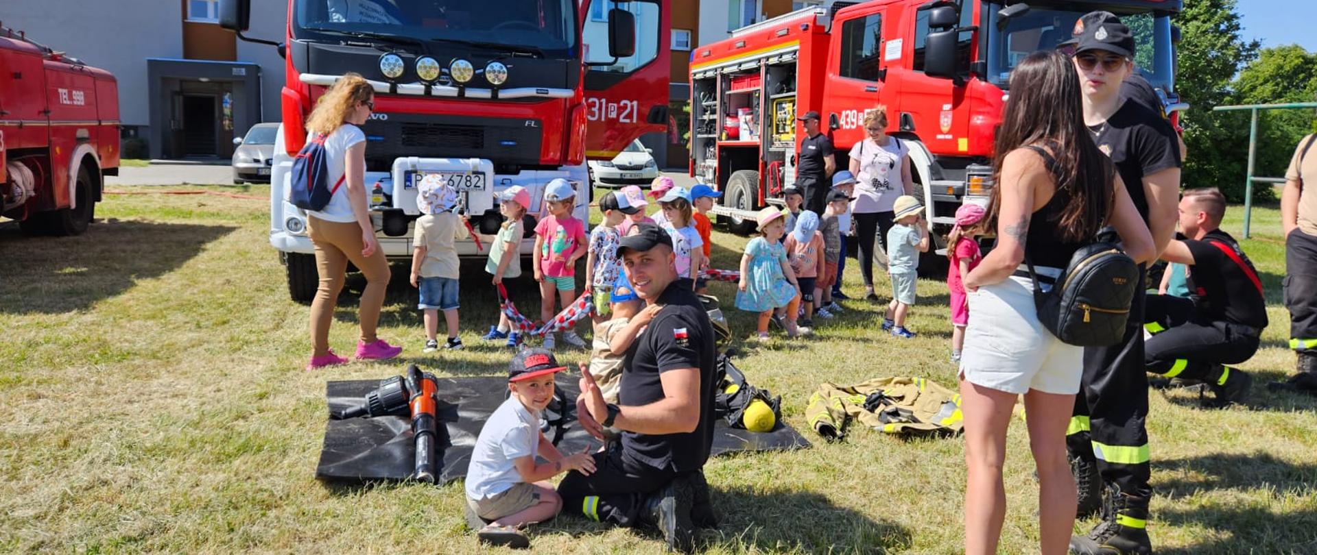 Zdjęcie przedstawia Strażaków którzy pokazują dzieciom sprzęt będący na wyposażeniu wozów strażackich. W tle znajdują się wozy strażackie