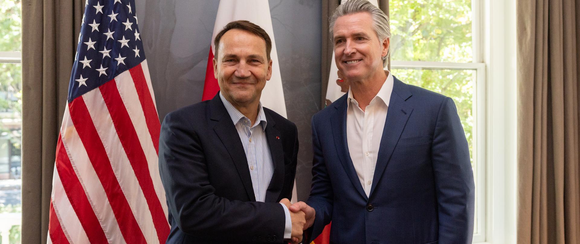 The photo shows two men in suits. Behind them is a window and three flags: the U.S., Poland, and California