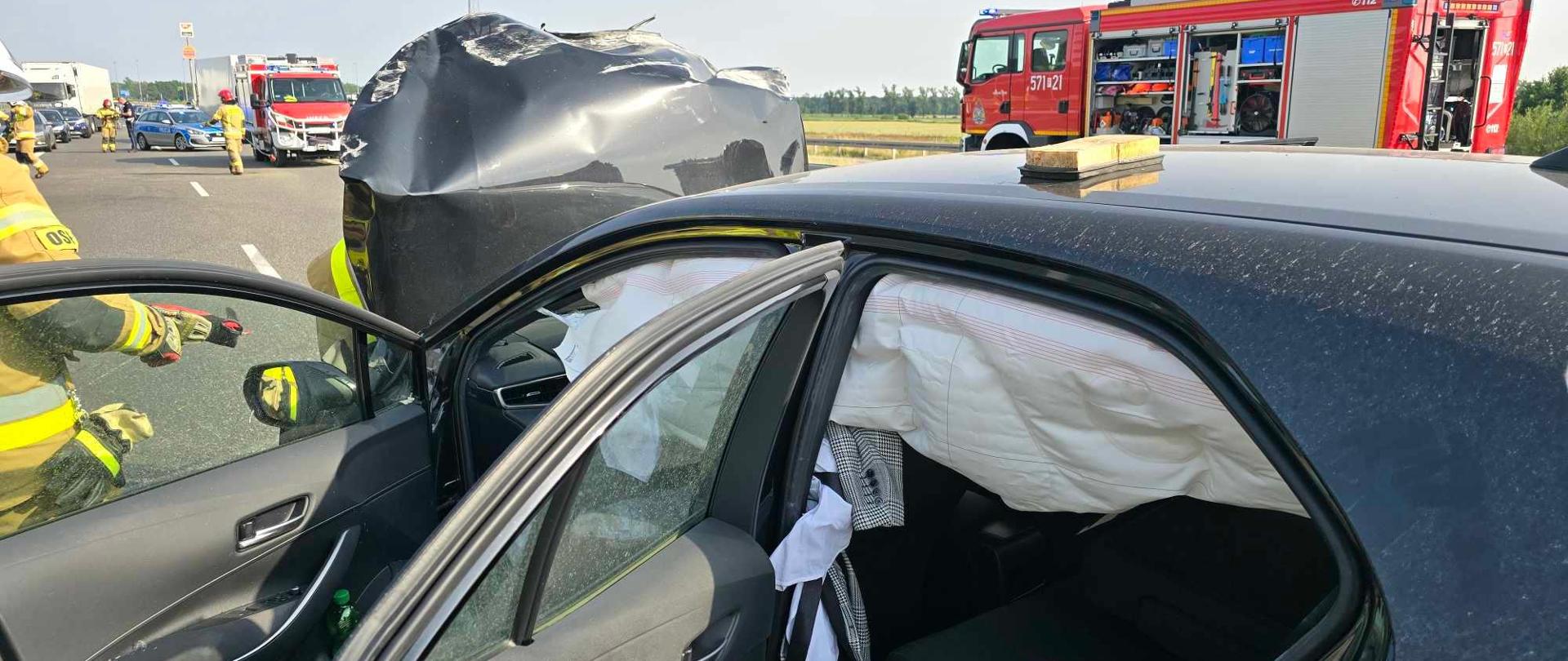 Na pierwszym planie widoczny jest rozbity czarny samochód osobowy. Drzwi z lewej strony są otwarte. Zadzialała kurtyna powietrzna.. Z lewej strony przy pojeździe stoi strażak, z prawej - wóz strażacki. Z przodu radiowóz policyjny i kolejne auto strażackie. Po jezdni rzemieszczają sie strażacy i policjant. Widać też stojące na pasie awaryjnym samochody cywilne. 