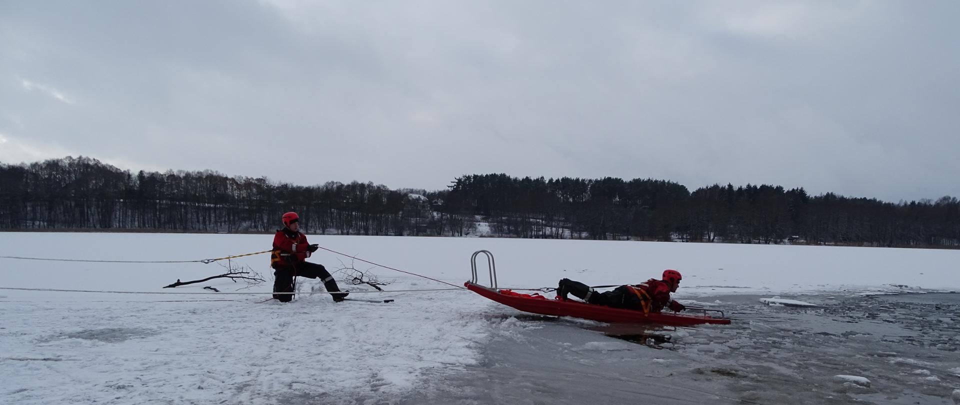 Zdjęcie wykonano podczas ćwiczeń ratownictwa lodowego strażaków z JRG Sępólno krajeńskie na nowej plaży w Sępólnie Krajeńskim. Przedstawia strażaka ubranego w kombinezon niezatapialny w kamizelce ratowniczej siedzącego na desce lodowej, zbliżającego się do pozoranta również ubranego kombinezon niezatapialny, unoszącego się na powierzchni wody po tym jak załamał się pod nim lód.