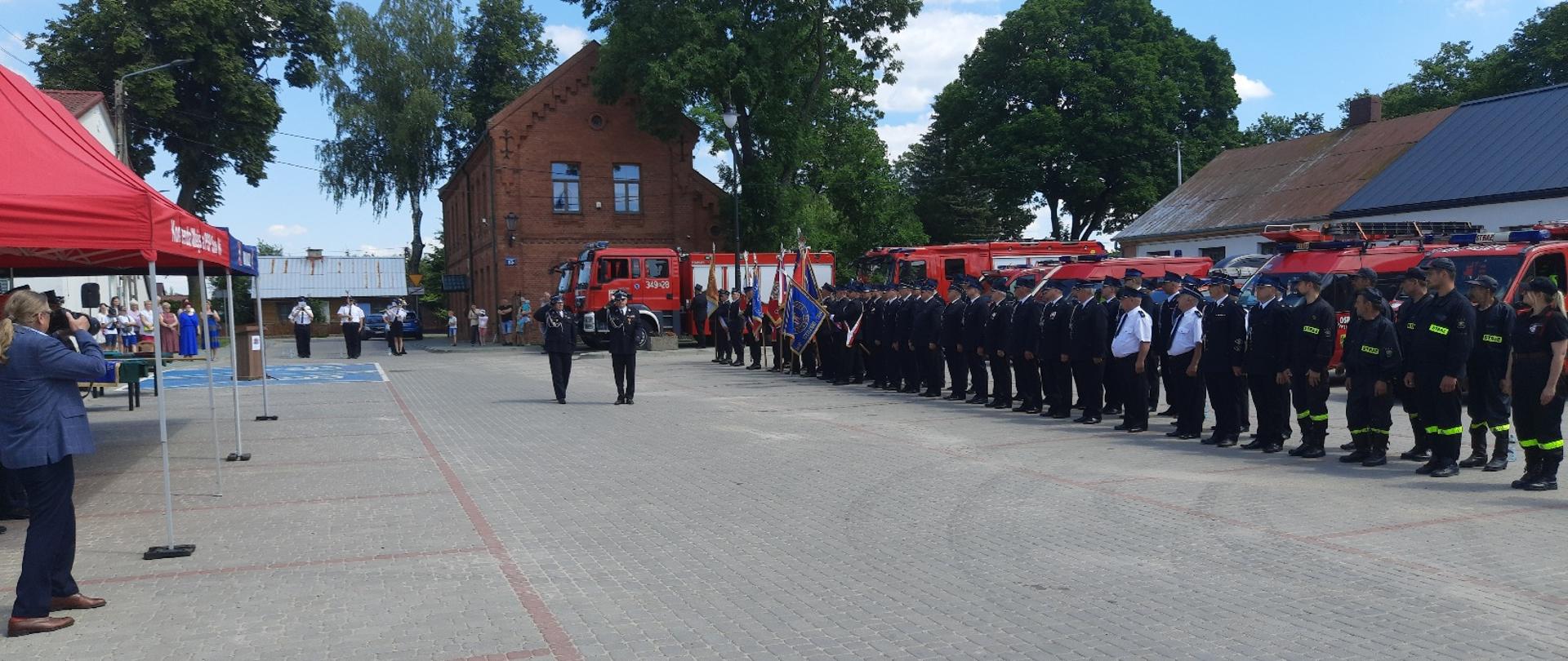 Zdjęcie obrazuje sztandary strażaków OSP i inne osoby w tle pojazdy pożarnicze i budynki