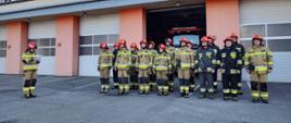Na zdjęciu widoczni są strażacy Jednostki Ratowniczo-Gaśniczej nr 1 w Tarnowie umundurowani w ubrania bojowe, stojący w dwuszeregu podczas apelu upamiętniającego strażaków z Ukrainy. W tle widoczny jest budynek KM PSP w Tarnowie oraz samochód bojowy stojący w bramie garażu.