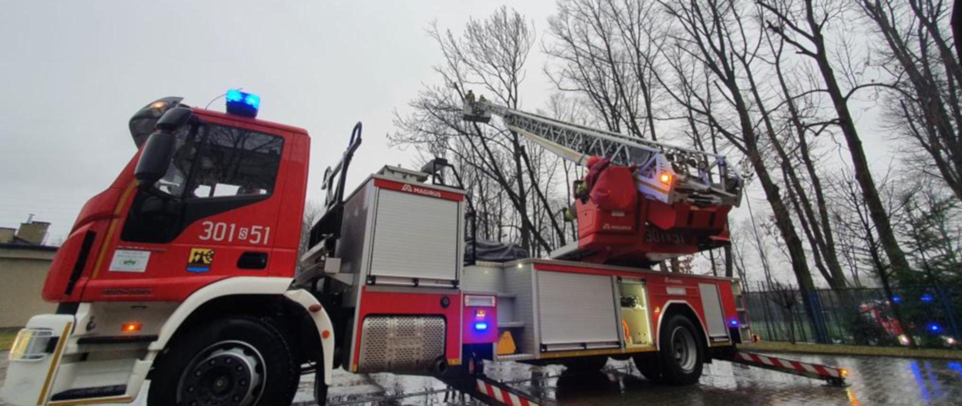 Na pierwszym planie samochód specjalny drabina z wysuniętym koszem, w którym znajduje się strażak. Ratownik piłą obcina gałęzie na uszkodzonym przez wiatr drzewie.