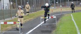 XI Powiatowe Zawody Sportowo-Pożarnicze OSP i MDP. Stadion w Miejskiej Górce. Na bieżni rozgrwana jest sztafeta pożarnicza 7x50 m z przeszkodami. Zawodnicy startujący na dwóch torach znajduja się w okolicach płotka lekkoatletycznego. 