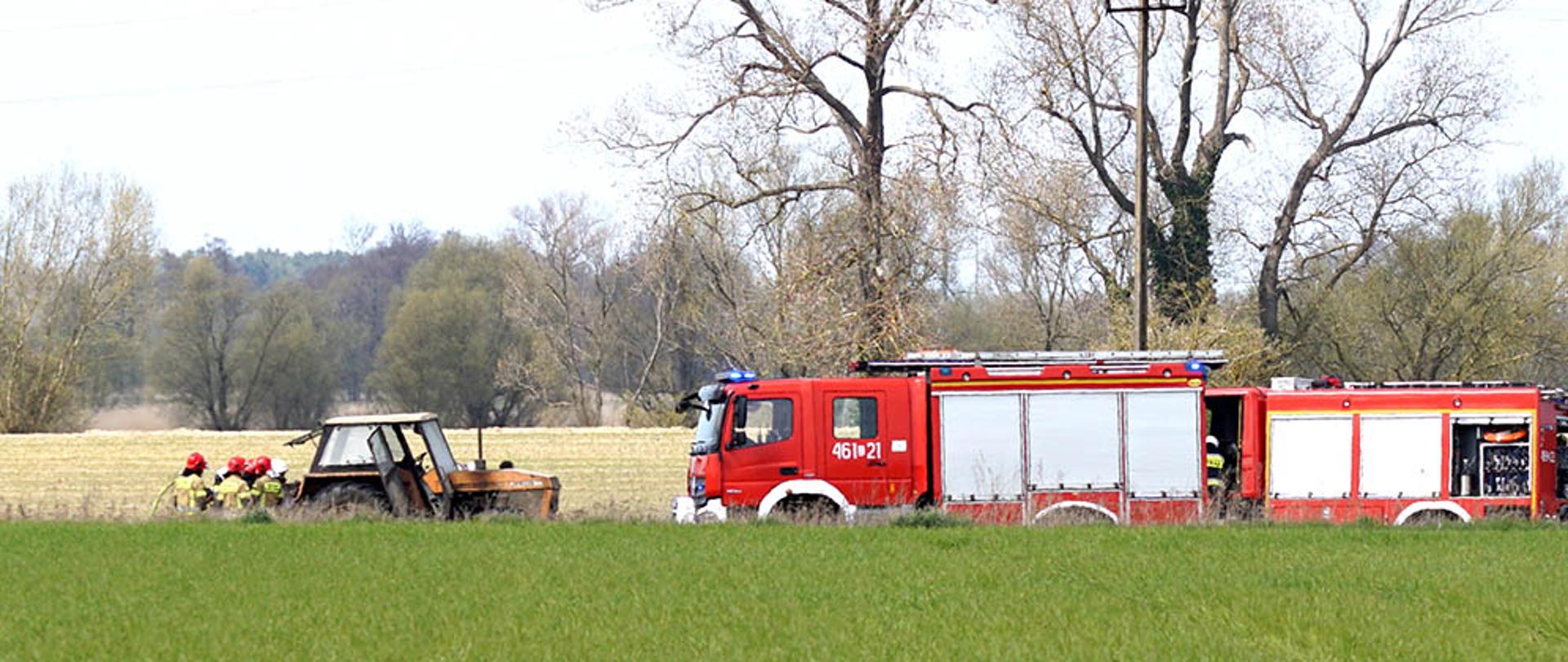 Zdjęcie przedstawia działania gaśnicze na polu. Widać strażaków, ciągnik oraz dwa wozy strażackie.