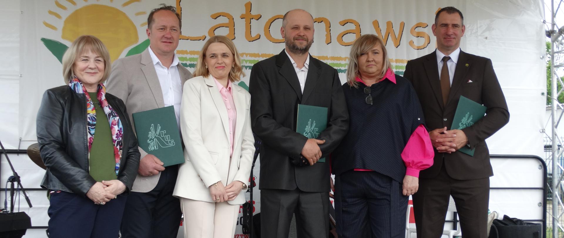 Trzy kobiety i trzech mężczyzn w odświętnych ubraniach stoją na scenie z napisem "Lato na wsi" i pozują do zdjęcia. Mężczyźni trzymają w rękach zielone ozdobne teczki ze złotym logo KRUS.