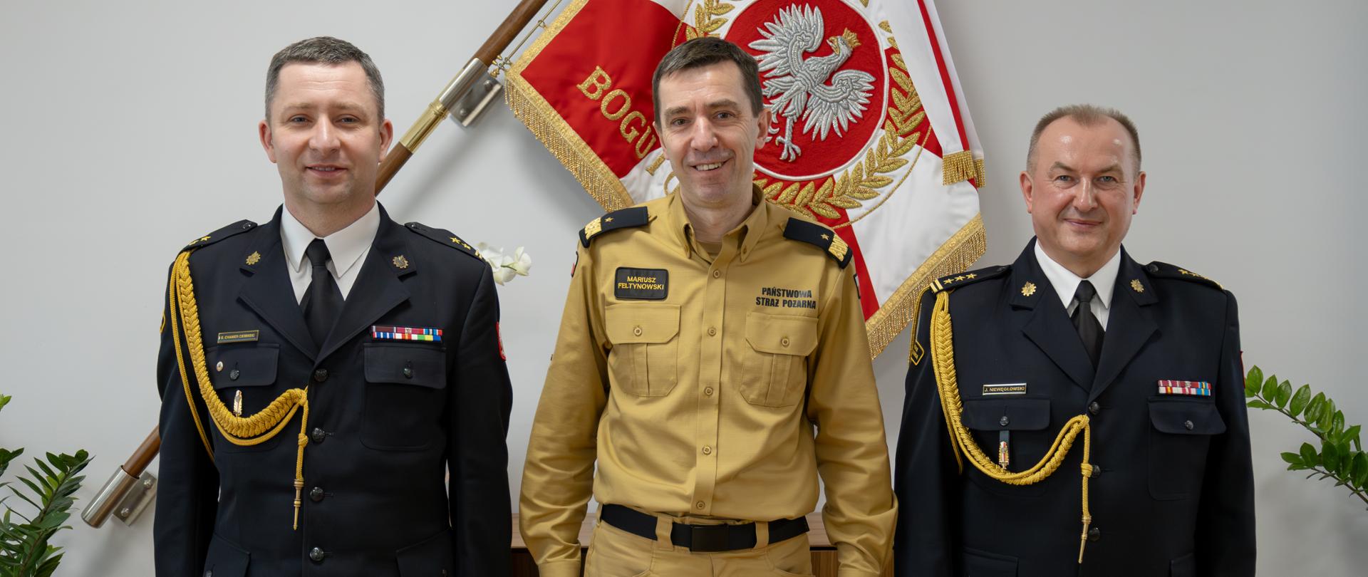 Strażak w mundurze w kolorze musztardowym stoi pomiędzy dwoma strażakami w mundurach wyjściowych ze sznurem za nimi na ścianie wisi sztandar Państwowej Straży Pożarnej oraz umieszczona jest komoda.