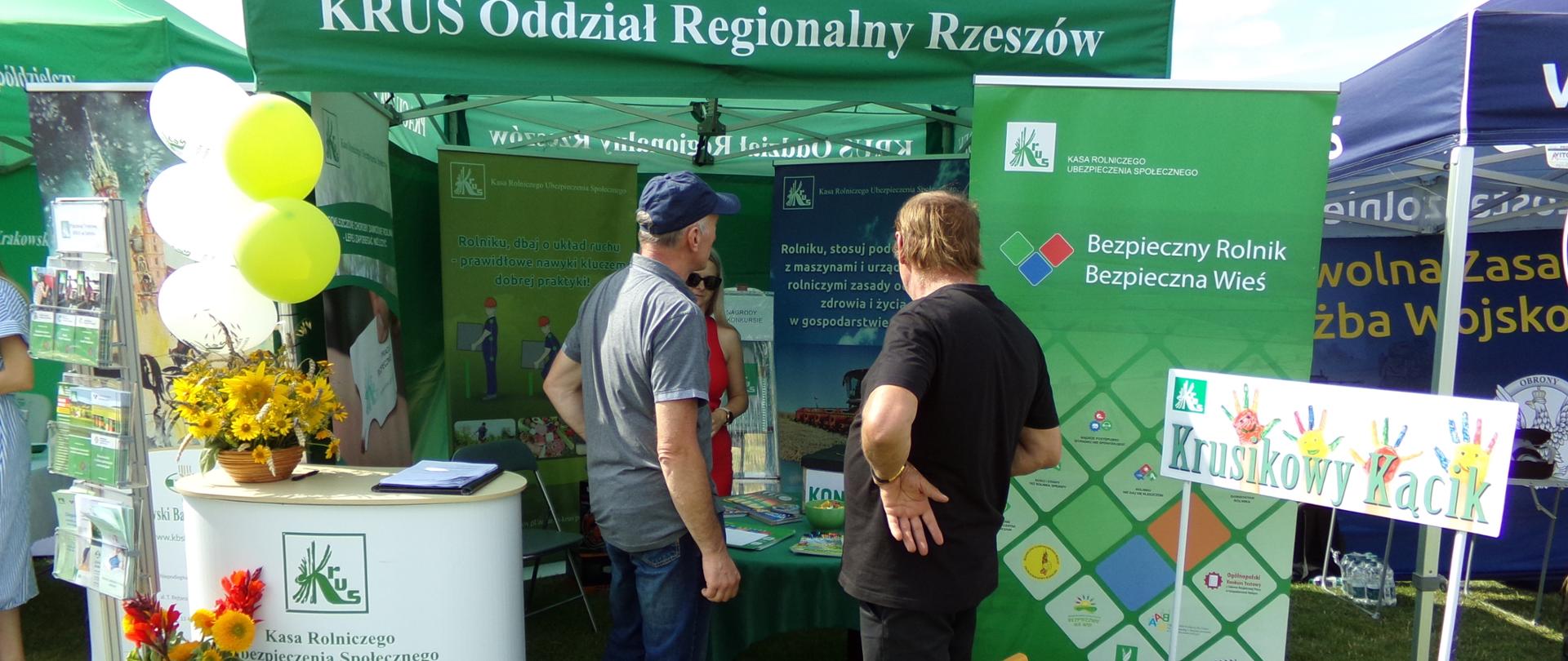 Przy stoisku informacyjno-prewencyjnym KRUS stoją dwaj mężczyźni, którzy z uwagą słuchają informacji udzielanych przez pracownika Kasy. Po ich lewej stronie znajduje się stolik z materiałami prewencyjnymi Kasy, a po prawej „Krusikowy Kącik” dla dzieci.
