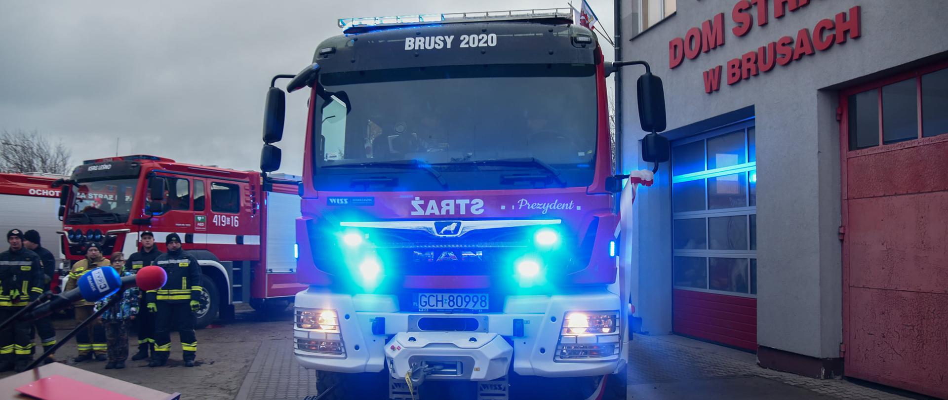 Przekazany samochód pożarniczy na tle budynku remizy. Widoczne włączone sygnały świetlne w kolorze niebieskim. W kadrze widoczna mównica a w tle strażacy stojący w szyku. 