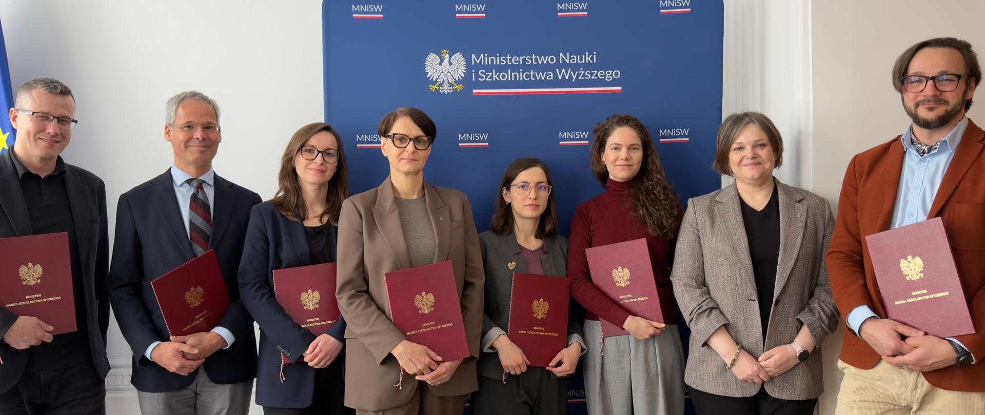 Zdjęcie zbiorowe, pod stojącym na tle białej ściany niebieskim banerem z napisem Ministerstwo Nauki i Szkolnictwa Wyższego stoi osiem osób, trzymają czerwone teczki, druga od prawej wiceminister Zioło-Pużuk w szarej marynarce.