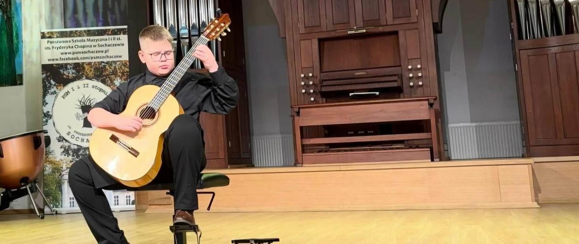 Na krześle siedzi chłopiec i gra na gitarze. Chłopiec ubrany jest w czarną koszulę i czarne spodnie oraz brązowe buty. Za nim w tle stoją organy muzyczne. Nad nim na ekranie wyświetlona jest grafika gitary i niebieski napis dwunasty Mały Festiwal Gitarowy 2 marca 2026 r.