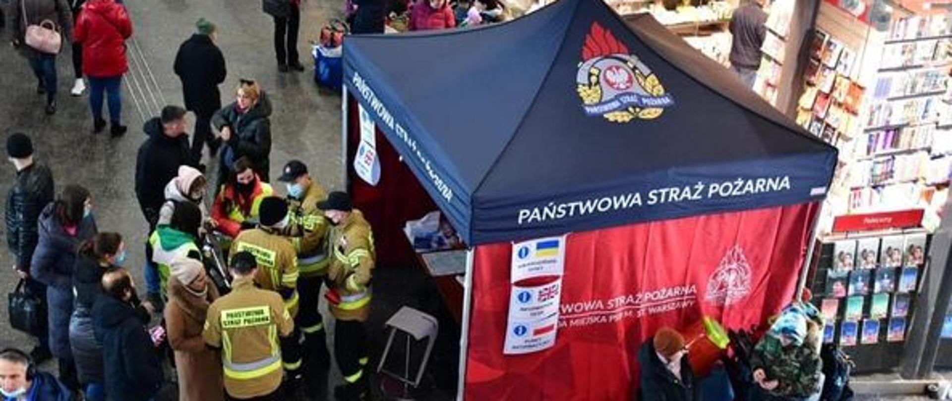 Zdjęcie przedstawia namiot Państwowej Straży Pożarnej wokół, którego zgromadzeni są strażacy oraz ludzie, którym strażacy udzielają niezbędnej pomocy