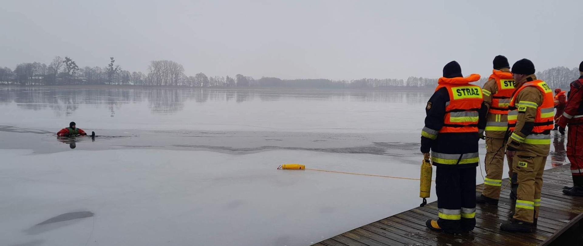 Po prawej stronie stronie zdjęcia na pomoście stoją tyłem strażacy ubrani w ubrania specjalne i kamizelki ratunkowe. Jeden z nich trzyma rzutkę. Po lewej na jeziorze na którym jest lód w przeręblu znajduje się strażak ubrany w suchy skafander. w połowie odległości między nimi widać leżącą na lodzie rzutkę.