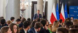 Na sali wypełnionej siedzącymi za stołami ludźmi za mównicą stoi minister Wieczorek i mówi do mikrofonu, za nim kolumny, z boku flagi Polski i UE.