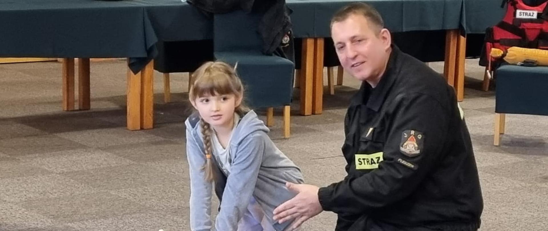 Strażak i kilkuletnia dziewczynka klęczą przy manekinie do ćwiczenia pierwszej pomocy