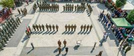 Zdjęcie wykonane z góry przedstawia płytę Rynku, na której odbywa się zbiórka żołnierska. Funkcjonariusze w mundurach typu moro stoją w szeregach. Za nimi dookoła stoją osoby cywilne i zaproszeni goście.