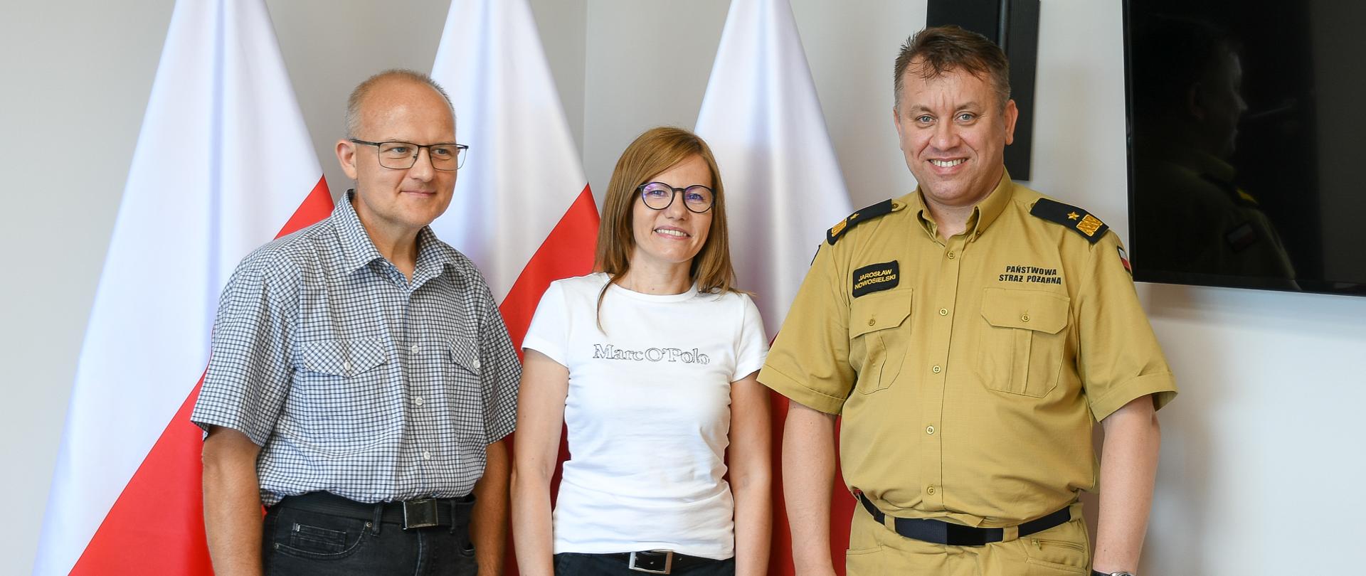 Na zdjęciu widać trzy osoby. W tle trzy flagi Polski.