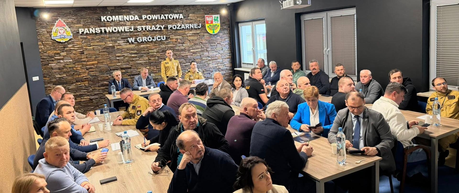 W sali konferencyjnej siedzi większa grupa osób przy długich stołach. Większość to mężczyźni, ale widać też kilka kobiet. Uczestnicy słuchają wystąpienia – niektórzy robią notatki lub trzymają telefony. Na ścianie widnieje napis „Komenda Powiatowa Państwowej Straży Pożarnej w Grójcu”. Pomieszczenie jest nowoczesne, z jasnym oświetleniem i projektorem pod sufitem.