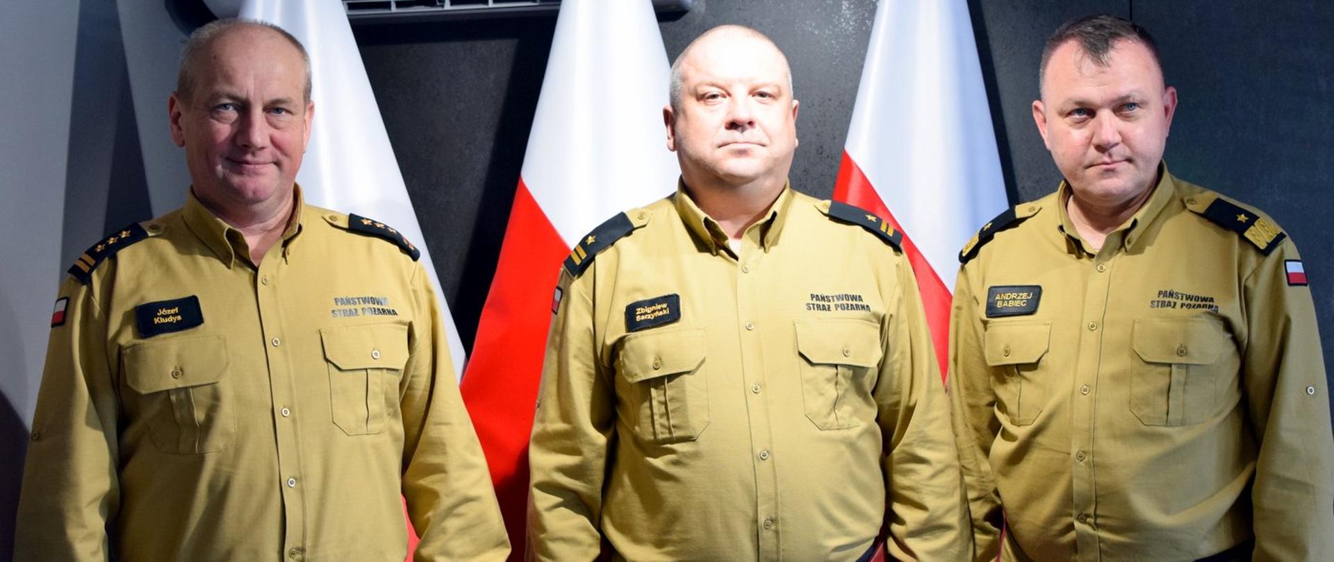 Zdjęcie zrobione wewnątrz pomieszczenia. Na zdjęciu trzech oficerów Państwowej Straży Pożarnej. Za nimi ustawione są biało-czerwone flagi.