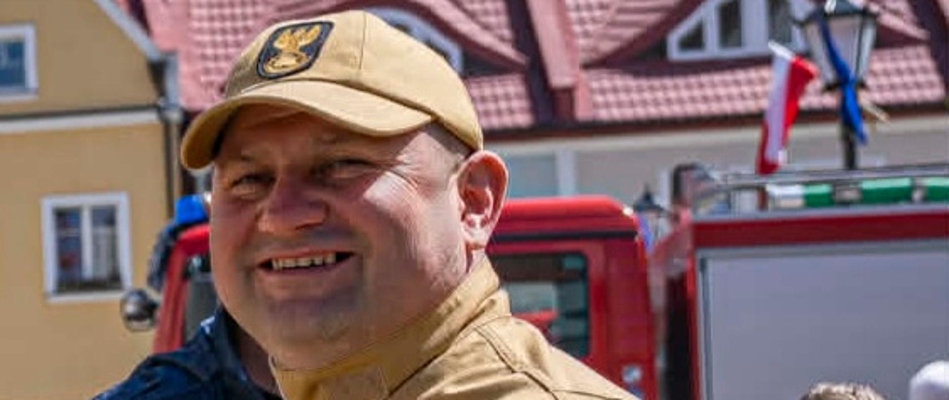 Zdjęcie przedstawia uśmiechniętego strażaka na pierwszym planie podczas plenerowego wydarzenia na miejskim rynku w słoneczny dzień.
Główna postać
Mundurowy: Mężczyzna ma na sobie piaskowy (beżowy) mundur służbowy Państwowej Straży Pożarnej oraz czapkę z daszkiem z godłem Polski.
Oznaczenia: Na piersi widoczna jest naszywka ze stopniem służbowym z trzema gwiazdkami (starszy kapitan). Na lewym ramieniu znajduje się flaga Polski oraz okrągła tarcza z emblematem PSP.
Mina: Mężczyzna uśmiecha się pogodnie, patrząc nieco w bok od obiektywu.
Tło i otoczenie
Architektura: W tle widoczne są budynki miejskiej zabudowy z charakterystycznym, czerwonym dachem krytym dachówką.
Pojazd i otoczenie: Za strażakiem stoi czerwony wóz strażacki oraz znajdują się inni uczestnicy wydarzenia.
Oznaczenie wodne: W prawym górnym rogu zdjęcia umieszczono logo i adres strony portalu internetowego „112 Polkowice”.