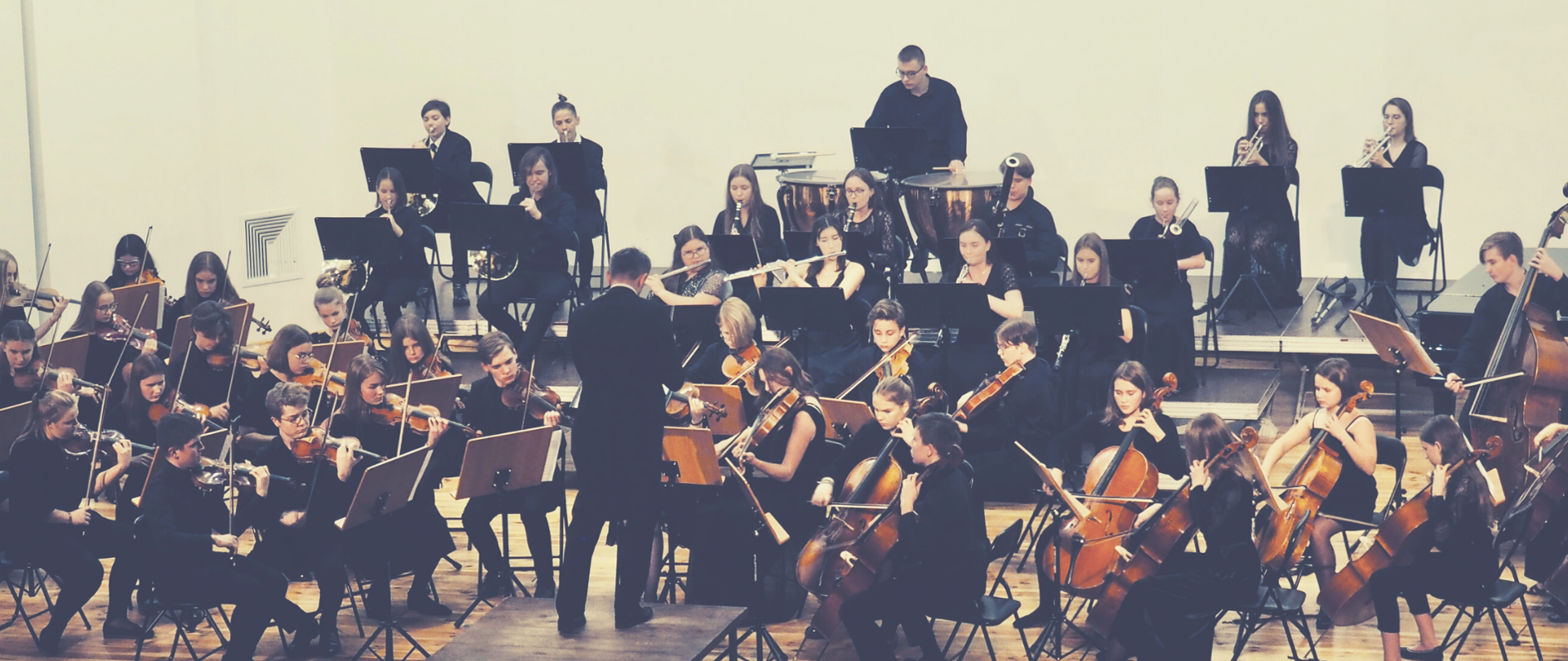 Orkiestra złożona z uczniów Państwowej Szkoły Muzycznej I i II stopnia imienia Karola Szymanowskiego w Płocku siedzi na scenie. Młodzi ludzie grają na instrumentach. Przed nimi dyrygent na podeście. Dyryguje.