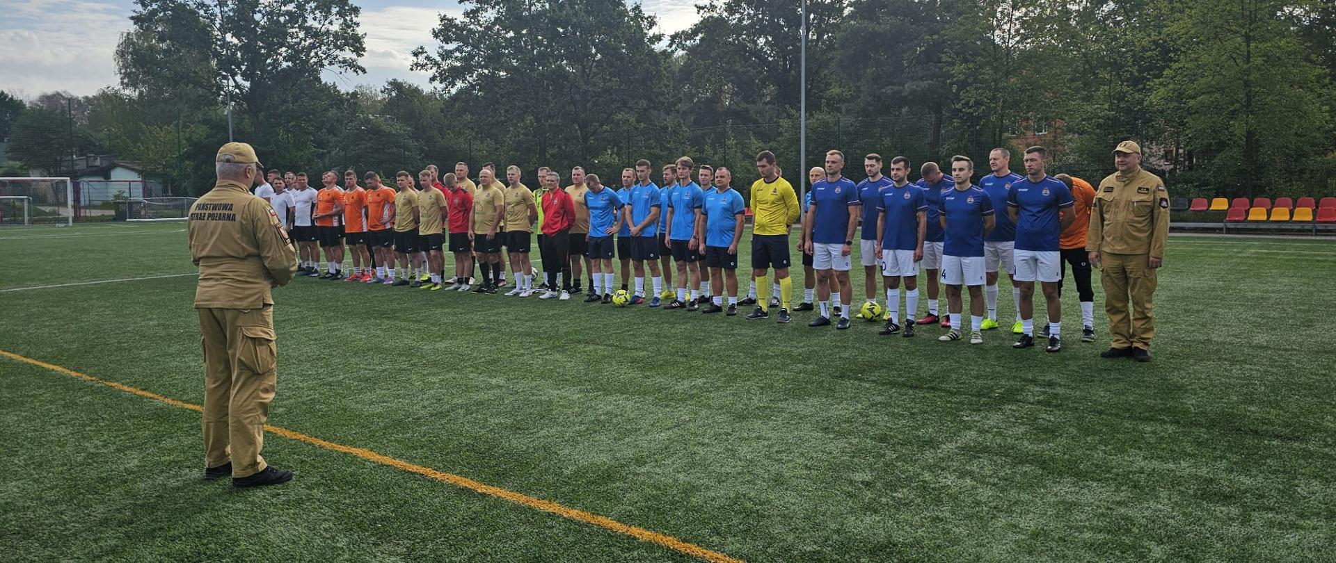 Na zdjęciu widać grupę strażaków Państwowej Straży Pożarnej podczas wydarzenia sportowego na boisku piłkarskim.
Na pierwszym planie, tyłem do aparatu, stoi funkcjonariusz w umundurowaniu Państwowej Straży Pożarnej, zwrócony w stronę uczestników. Na jego mundurze widoczny jest napis „PAŃSTWOWA STRAŻ POŻARNA”.
W głębi boiska ustawieni są w szeregu strażacy ubrani w sportowe stroje w różnych kolorach – m.in. pomarańczowych, beżowych, czerwonych, jasnoniebieskich i granatowych. Część zawodników ma przy sobie piłki, a jeden z uczestników występuje w stroju bramkarskim. Po prawej stronie znajduje się drugi funkcjonariusz PSP w umundurowaniu służbowym.
Całość odbywa się na pełnowymiarowym boisku ze sztuczną nawierzchnią, otoczonym ogrodzeniem i drzewami. W tle widoczne są również trybuny z kolorowymi siedziskami oraz infrastruktura sportowa.
