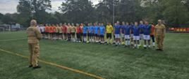 Na zdjęciu widać grupę strażaków Państwowej Straży Pożarnej podczas wydarzenia sportowego na boisku piłkarskim.
Na pierwszym planie, tyłem do aparatu, stoi funkcjonariusz w umundurowaniu Państwowej Straży Pożarnej, zwrócony w stronę uczestników. Na jego mundurze widoczny jest napis „PAŃSTWOWA STRAŻ POŻARNA”.
W głębi boiska ustawieni są w szeregu strażacy ubrani w sportowe stroje w różnych kolorach – m.in. pomarańczowych, beżowych, czerwonych, jasnoniebieskich i granatowych. Część zawodników ma przy sobie piłki, a jeden z uczestników występuje w stroju bramkarskim. Po prawej stronie znajduje się drugi funkcjonariusz PSP w umundurowaniu służbowym.
Całość odbywa się na pełnowymiarowym boisku ze sztuczną nawierzchnią, otoczonym ogrodzeniem i drzewami. W tle widoczne są również trybuny z kolorowymi siedziskami oraz infrastruktura sportowa.
