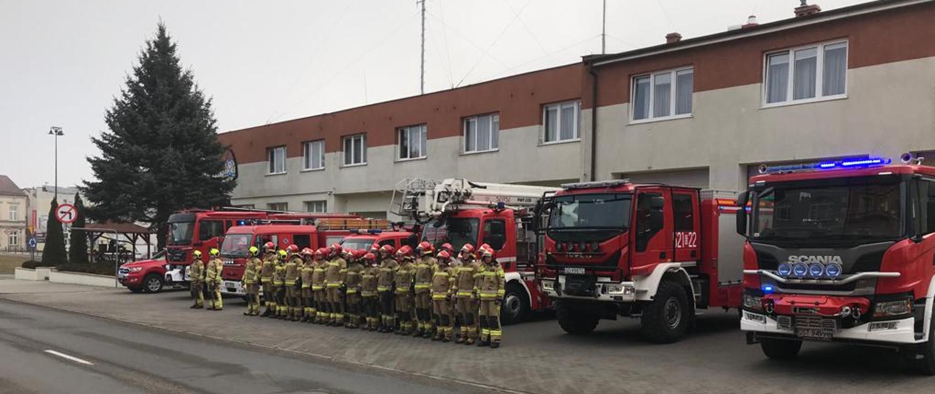 Zdjęcie przedstawia strażaków PSP Starogard Gdański oraz OSP Starogard Gdański przed Komendą na tle samochodów pożarniczych. Włączone sygnały świetlne