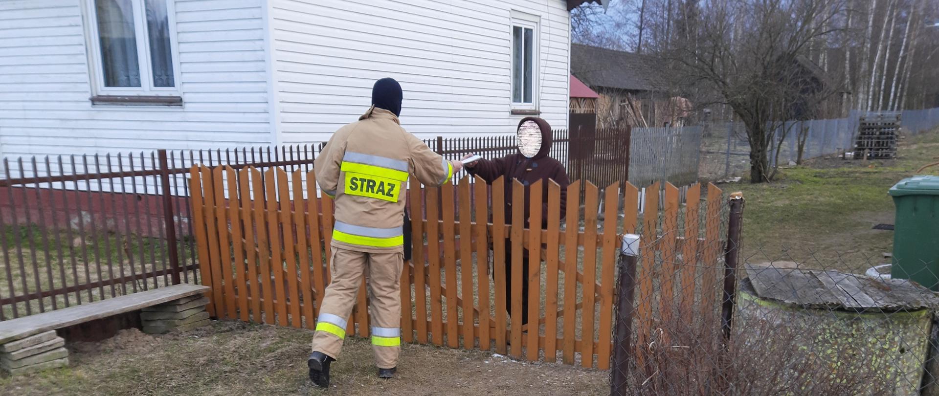 Strażak OSP podczas akcji dystrybucji ulotek informacyjnych na temat profilaktyki oraz szczepień przeciwko COVID-19. Strażak przekazuje ulotkę mieszkance jednej z miejscowości powiatu bielskiego.