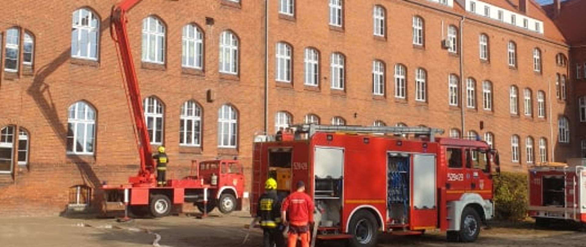 Zdjęcie przedstawia samochody pożarnicze na tle budynku szkoły.