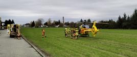Na pierwszym planie widoczny śmigłowiec LPR, który wylądował na polu. Obok niego strażacy wraz z Ratownikami. Po lewej stronie karetka pogotowia, która przywiozła poszkodowanego do śmigłowca. 