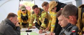 Narada funkcjonariuszy straży pożarnej w namiocie. Siedzą i stoją dookoła stołu. Na stole leżą mapa, dokumenty i długopisy, oraz stoi laptop.
