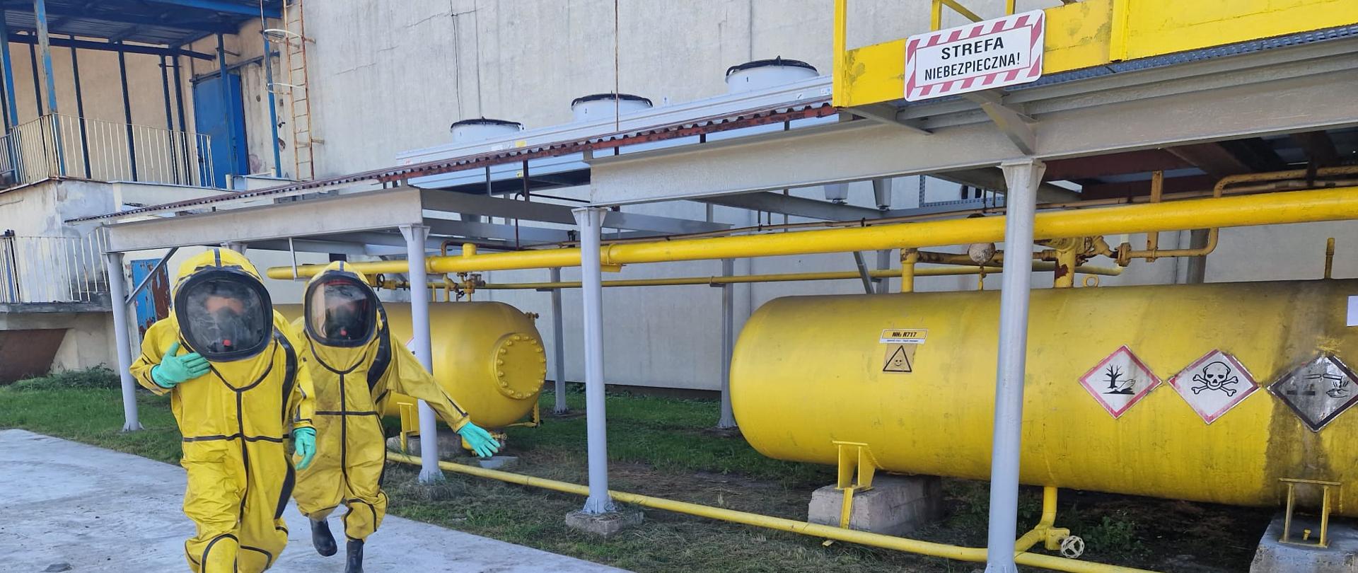 Na zdjęciu duże zbiorniki w kształcie walca koloru żółtego, na nich widoczne oznakowania informujące o różnego rodzaju zagrożeniach. Przy nich dwaj strażacy w ubraniach ochrony przeciwchemicznej.