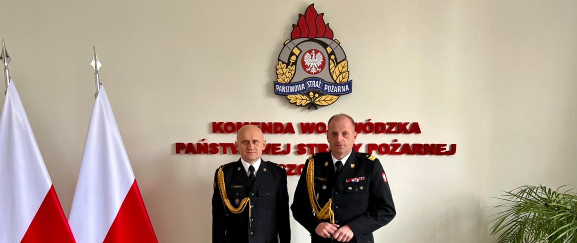 Na tle ścianki z logiem PSP i flagami stoi Komendant Wojewódzki i Komendant Powiatowy w mundurach galowych. 