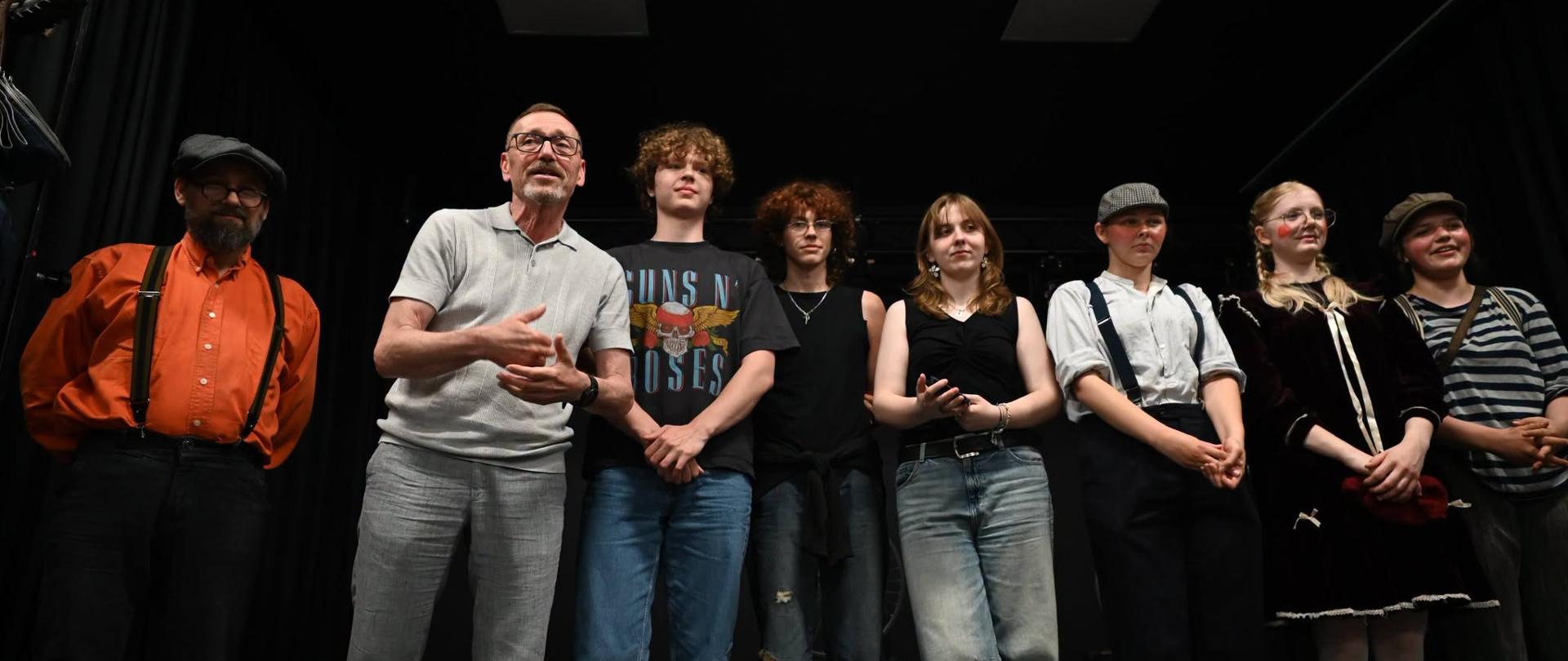 Na scenie na tle czarnej ściany stoją cztery dziewczyny z prawej strony (trzy w kostiumach teatralnych), pośrodku dwóch chłopców i dwóch mężczyzn z lewej strony