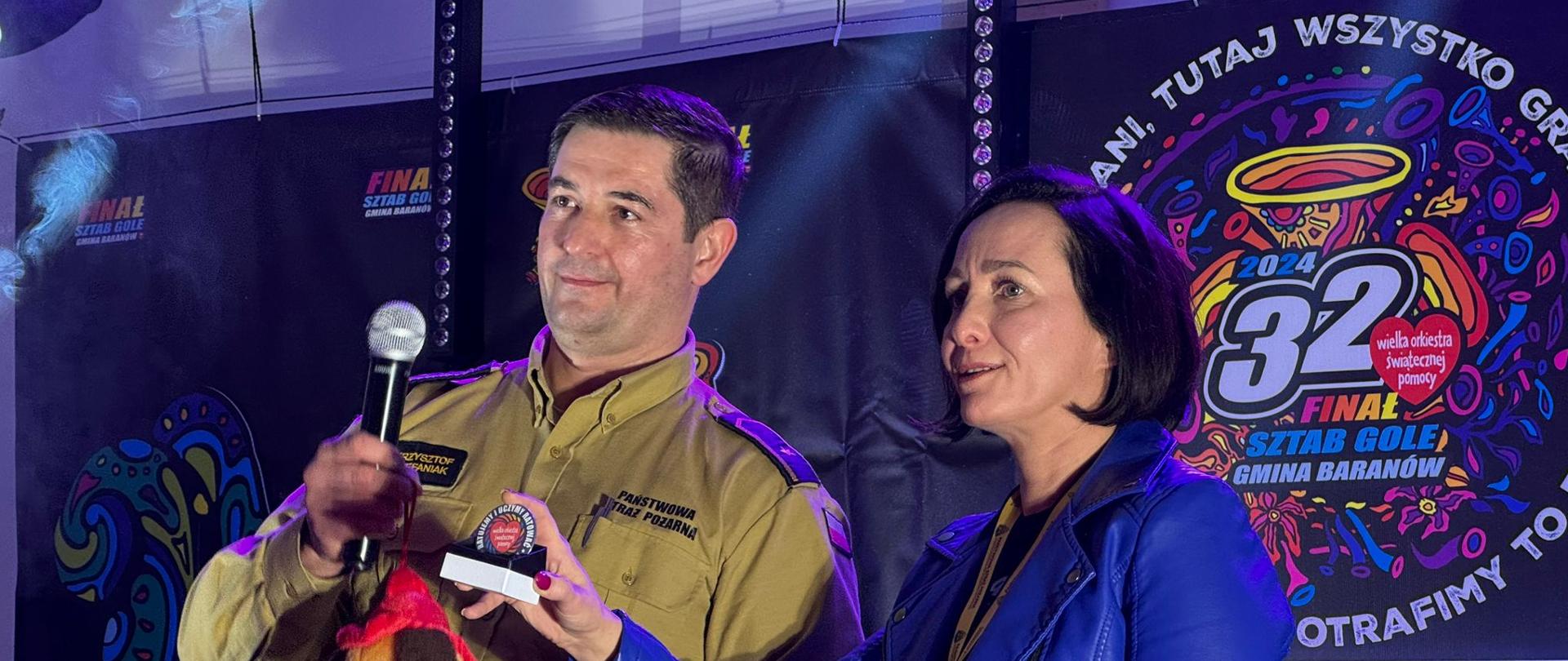 Licytacja strażackiej maskotki trzymanej przez asp. Krzysztofa Stefaniaka obok niego prowadząca.